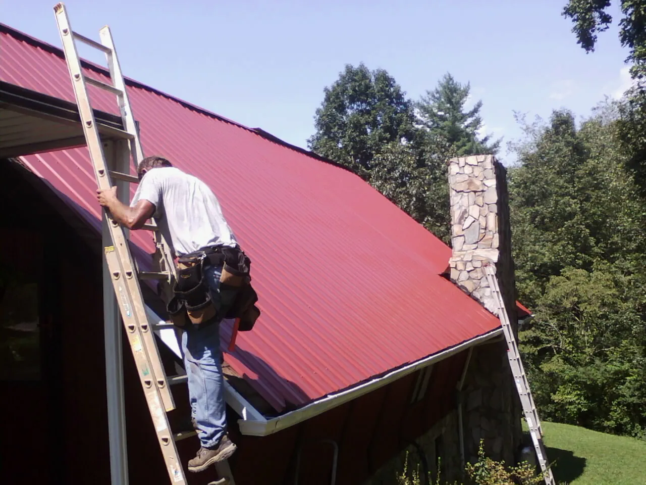 Roofer on a ladder beside a red metal roof and stone chimney — Soffit & Fascia Repair by Roofing Brothers in Big Clifty
